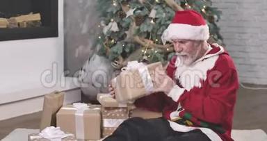 白种人，留着白胡子和胡子，拿着礼品盒，摇着它。 戴着红帽子和圣诞毛衣的圣诞老人